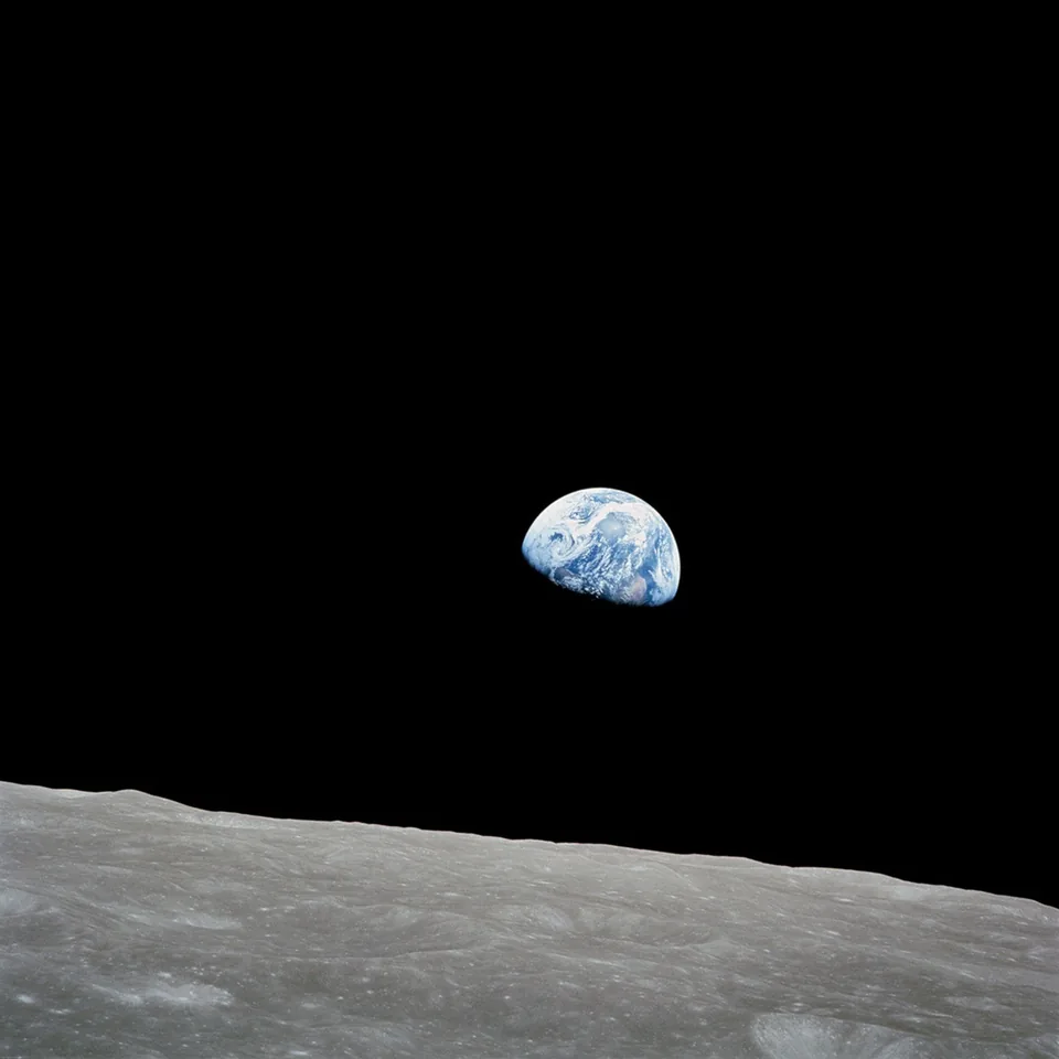 A small partly sunlit Earth rises above the cratered lunar horizon against black space.