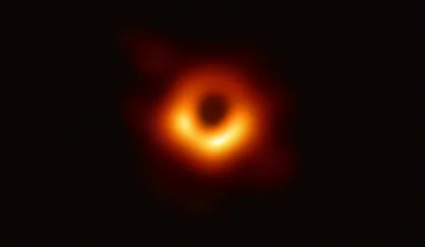 An uneven bright radio ring surrounds a dark centre, with the lower arc brighter than the upper arc.
